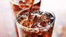 A tall glass is filled with ice cubes and fizzing brown cola. There's a small stream of brown soda being poured into the almost full glass of ice and soda.